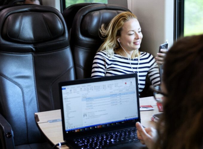 Icomera's IcoShape data traffic management solution offers many benefits for passengers and operational systems. This photo shows a female passenger on a train using a laptop in the foreground. Another female passenger wearing headphones and using a phone is sitting in the seat opposite
