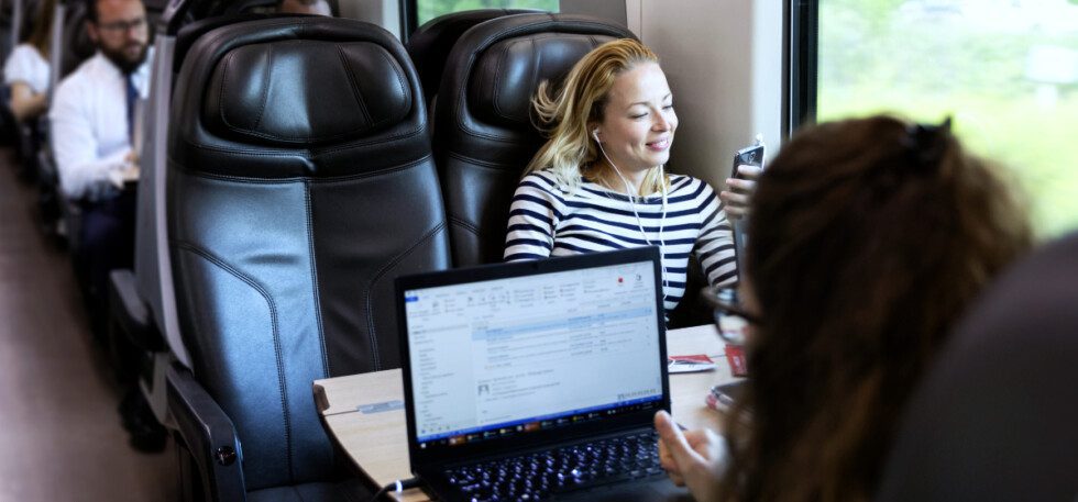 Icomera's IcoShape data traffic management solution offers many benefits for passengers and operational systems. This photo shows a female passenger on a train using a laptop in the foreground. Another female passenger wearing headphones and using a phone is sitting in the seat opposite