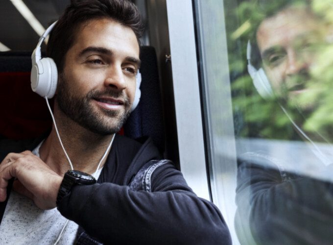 A male passenger sits on a train. He is listening to music through headphones and staring outside the window