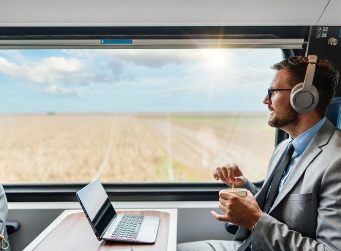 A male passenger wearing headphones works on a laptop while enjoying a coffee on his journey.