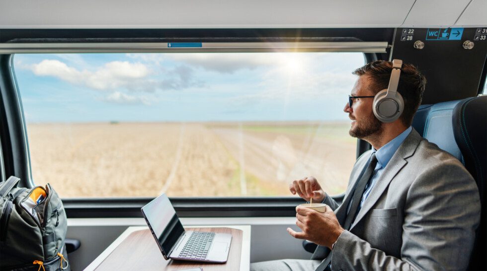 A male passenger wearing headphones works on a laptop while enjoying a coffee on his journey.