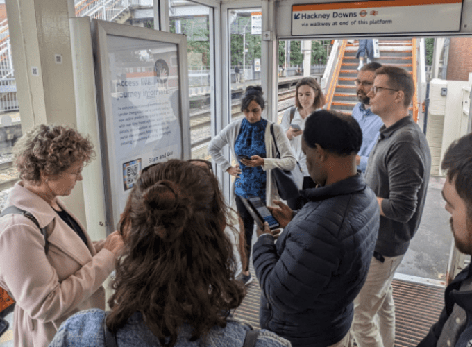 Luna users at Hackney Downs station, London. They are standing in front of an accessibility information sign designed to be used with Luna, testing the application on their phones.