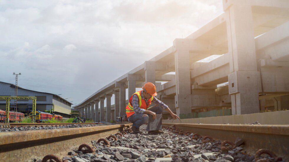 A railway engineer wearing orange high-visibility clothing and hard hat kneels down to inspect a train tack