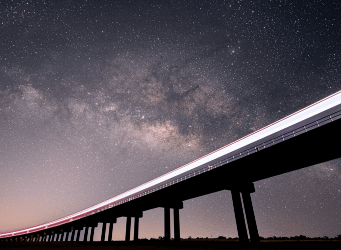 A light trail from a train stretches across a rail bridge, in front of a clear, starry night sky with the Milky Way spiral arm visible.