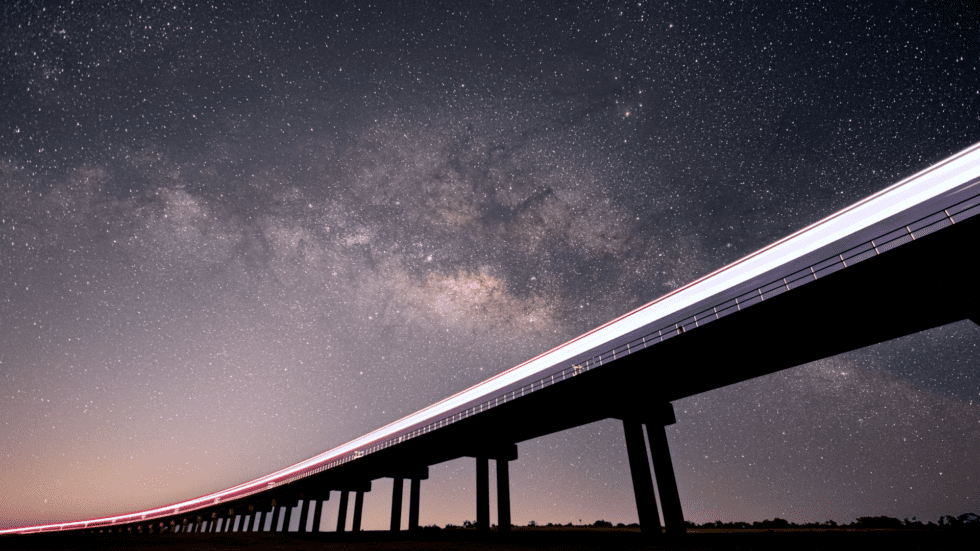 A light trail from a train stretches across a rail bridge, in front of a clear, starry night sky with the Milky Way spiral arm visible.